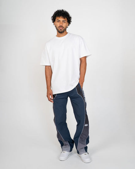 A man modeling a white crewneck t-shirt and navy blue athletic pants with gray side panels and white piping, paired with white sneakers, against a light gray studio background.