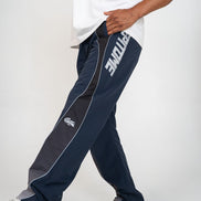 A person wearing navy blue track pants with a dark gray side panel, white piping, and a large white 'EPITOME' logo on the upper leg, paired with a white t-shirt and white sneakers against a light gray studio background.