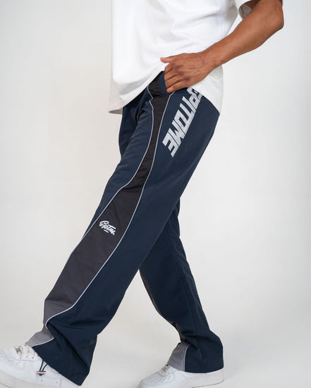 A person wearing navy blue track pants with a dark gray side panel, white piping, and a large white 'EPITOME' logo on the upper leg, paired with a white t-shirt and white sneakers against a light gray studio background.