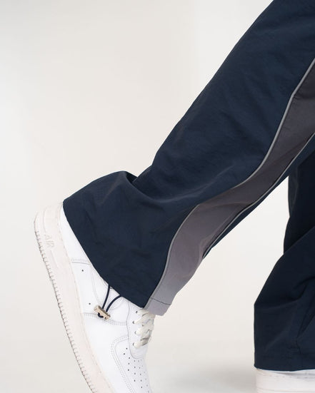 Close-up of a person's leg wearing navy blue pants with a gray side stripe and a white sneaker, shown against a plain light background.