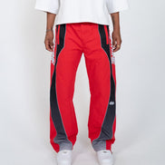 A person wearing red track pants with bold black side panels and white sneakers, photographed against a clean light gray studio background.
