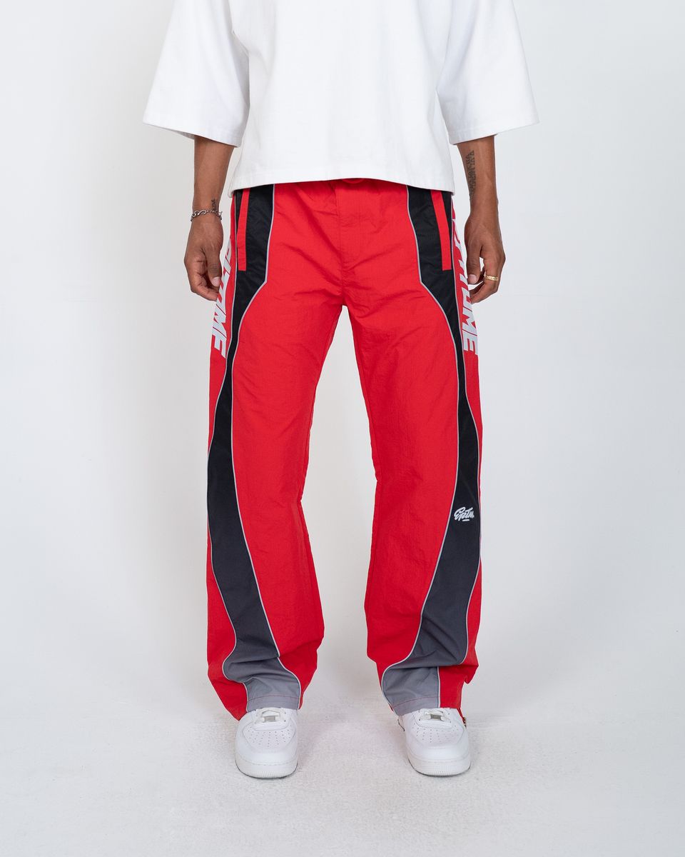 A person wearing red track pants with bold black side panels and white sneakers, photographed against a clean light gray studio background.