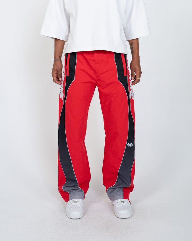 A person wearing red track pants with bold black side panels and white sneakers, photographed against a clean light gray studio background.