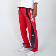 A person wearing red track pants with black side panels and white 'EPITOME' branding, paired with a white t-shirt and white sneakers, photographed against a light gray studio background.