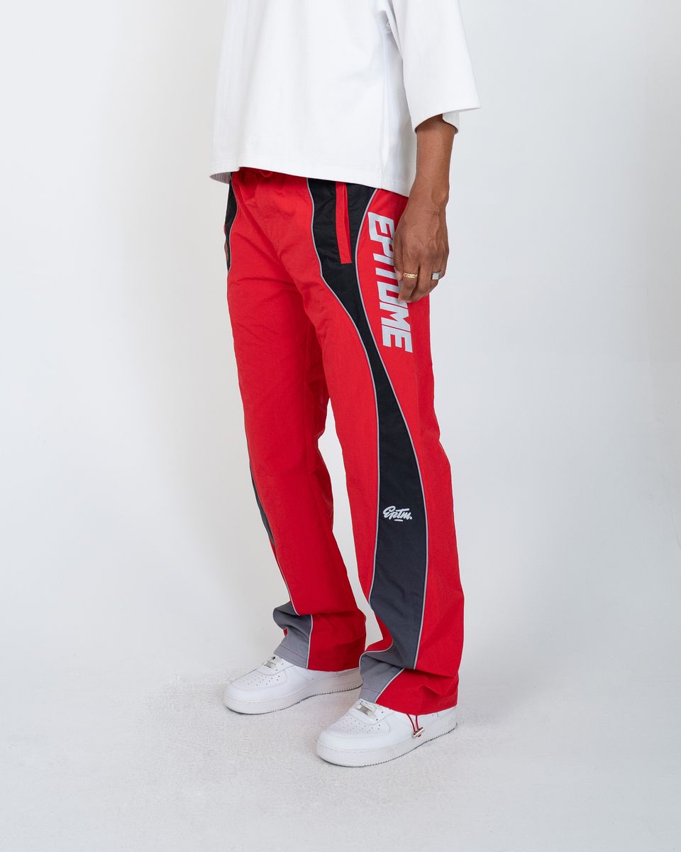 A person wearing red track pants with black side panels and white 'EPITOME' branding, paired with a white t-shirt and white sneakers, photographed against a light gray studio background.