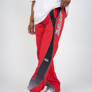 A person wearing red track pants with a black side panel, gray toe caps, and white sneakers against a light gray studio background.