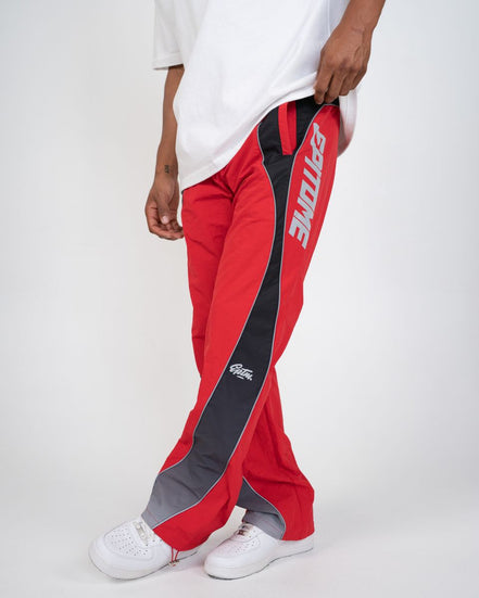 A person wearing red track pants with a black side panel, gray toe caps, and white sneakers against a light gray studio background.