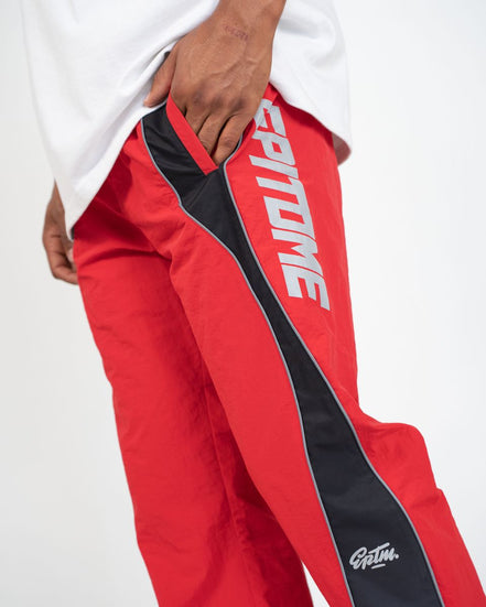 Red and black track pants with white 'EPITOME' text on the leg and a white 'Gatm.' logo near the hem, shown on a model wearing a white t-shirt against a white studio background.