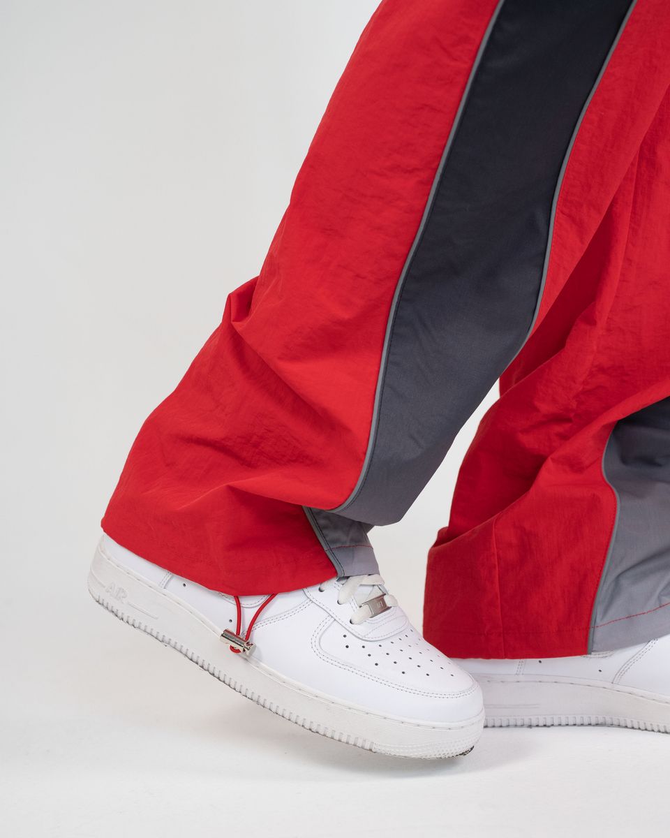 Close-up of a person's legs wearing red and dark gray wide-leg pants with white sneakers against a light gray studio background.