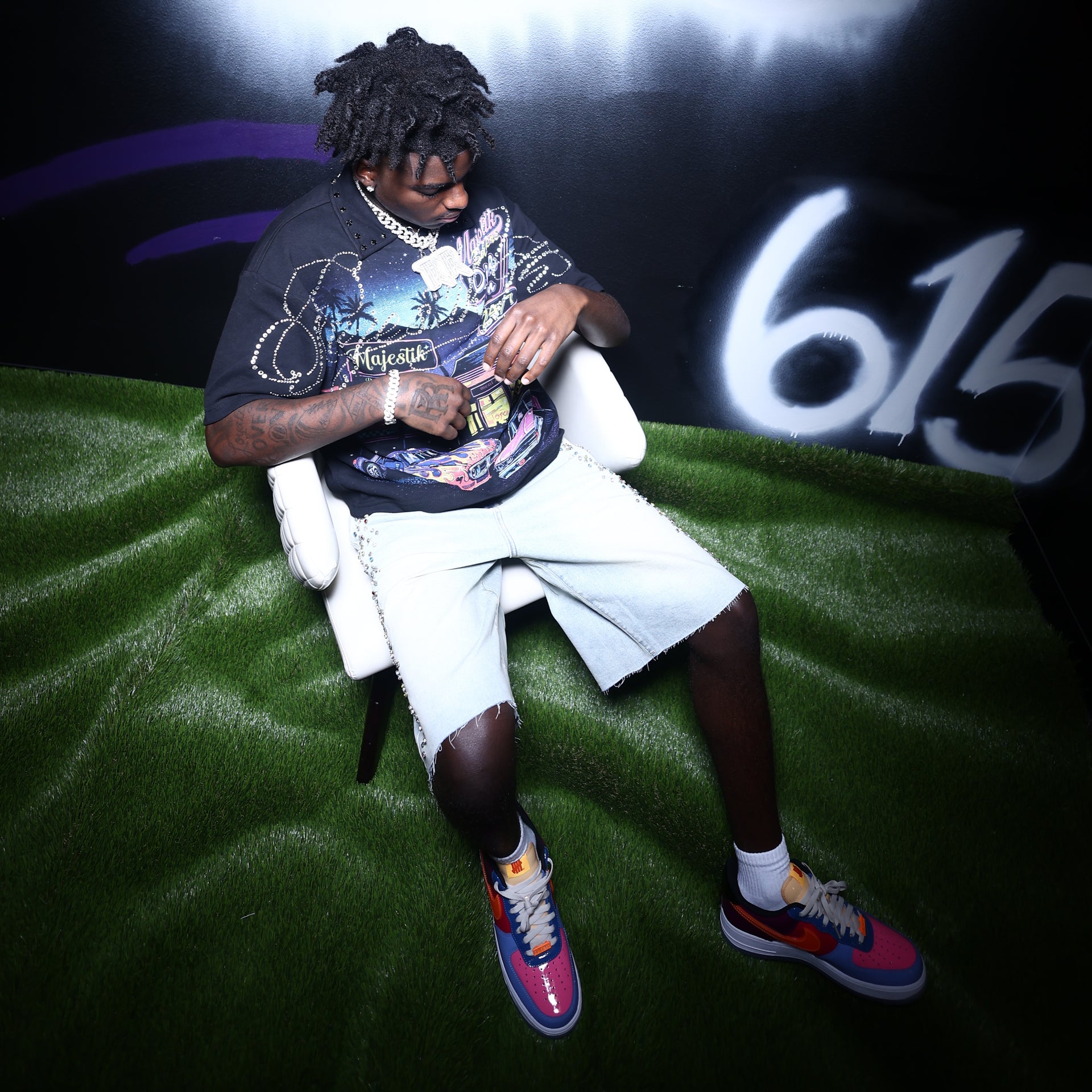 A man sits in a white armchair on a green turf-like surface, wearing a black graphic polo, light denim shorts, and colorful sneakers. The background is dark with a large white '615' graphic and purple streaks.