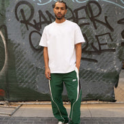 Person wearing a white t-shirt and green tracksuit with white stripes standing in front of a graffiti-covered wall.