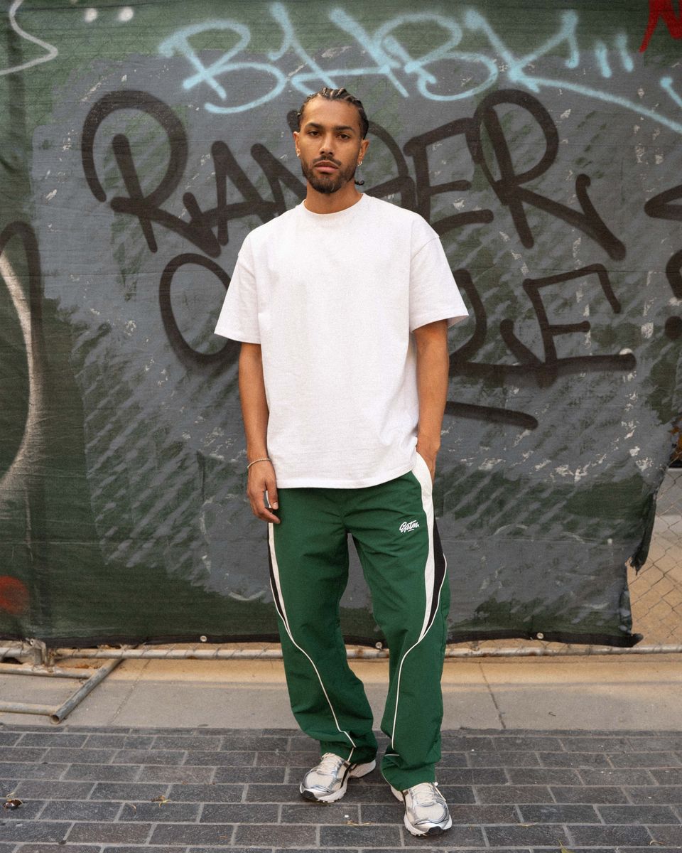 Person wearing a white t-shirt and green tracksuit with white stripes standing in front of a graffiti-covered wall.