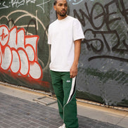 Man wearing a white t-shirt and green tracksuit with white stripes standing in front of a graffiti-covered wall.