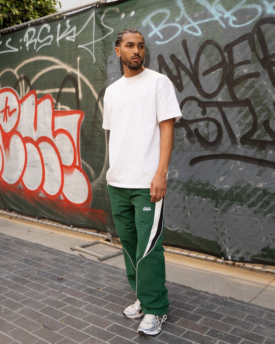 Man wearing a white t-shirt and green tracksuit with white stripes standing in front of a graffiti-covered wall.