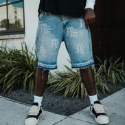 Person wearing light blue denim shorts with white text and black sneakers on a sidewalk.