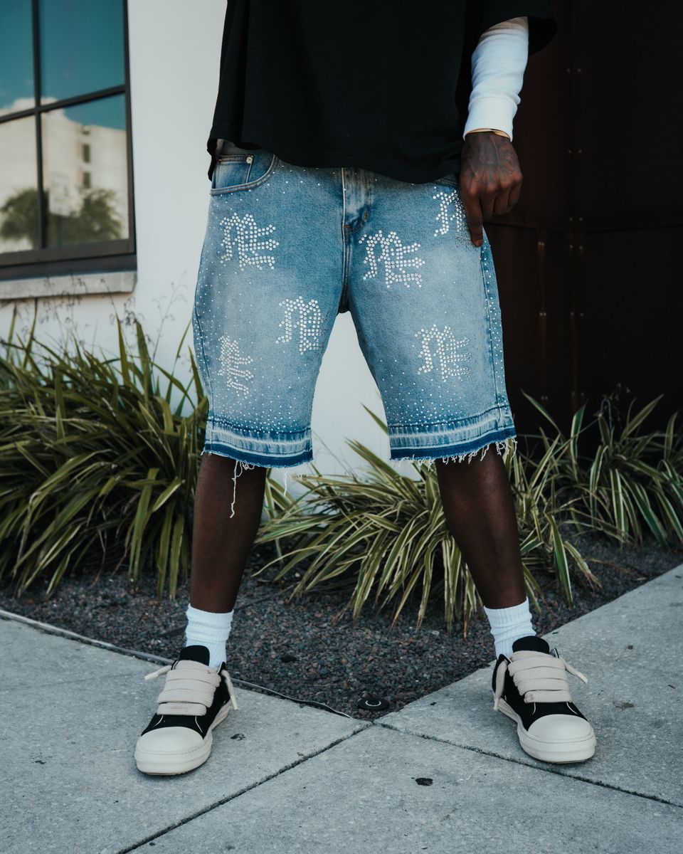Person wearing light blue denim shorts with white text and black sneakers on a sidewalk.