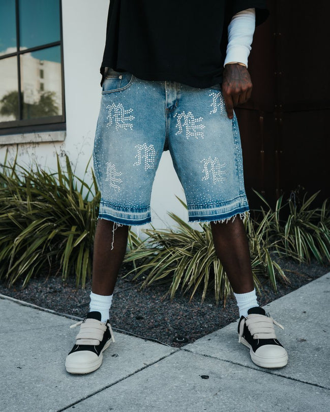 Person wearing light blue denim shorts with white text and black sneakers on a sidewalk.