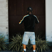 Person wearing a black hoodie with colorful stripes, denim shorts, and brown boots standing in front of a dark door.
