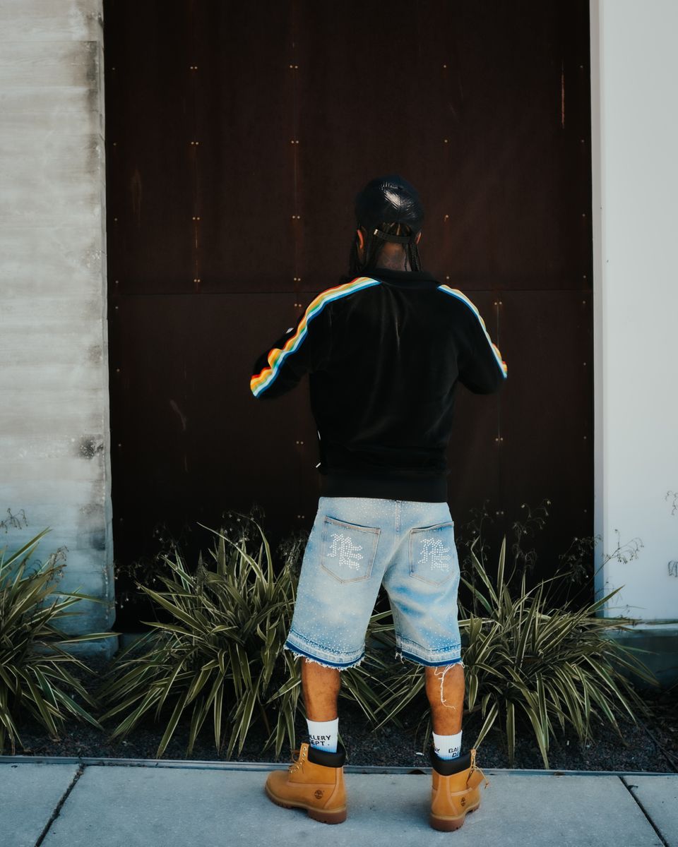 Person wearing a black hoodie with colorful stripes, denim shorts, and brown boots standing in front of a dark door.