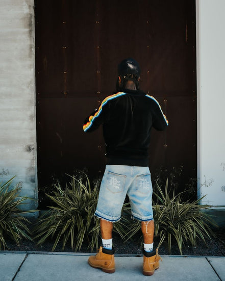 Person wearing a black hoodie with colorful stripes, denim shorts, and brown boots standing in front of a dark door.