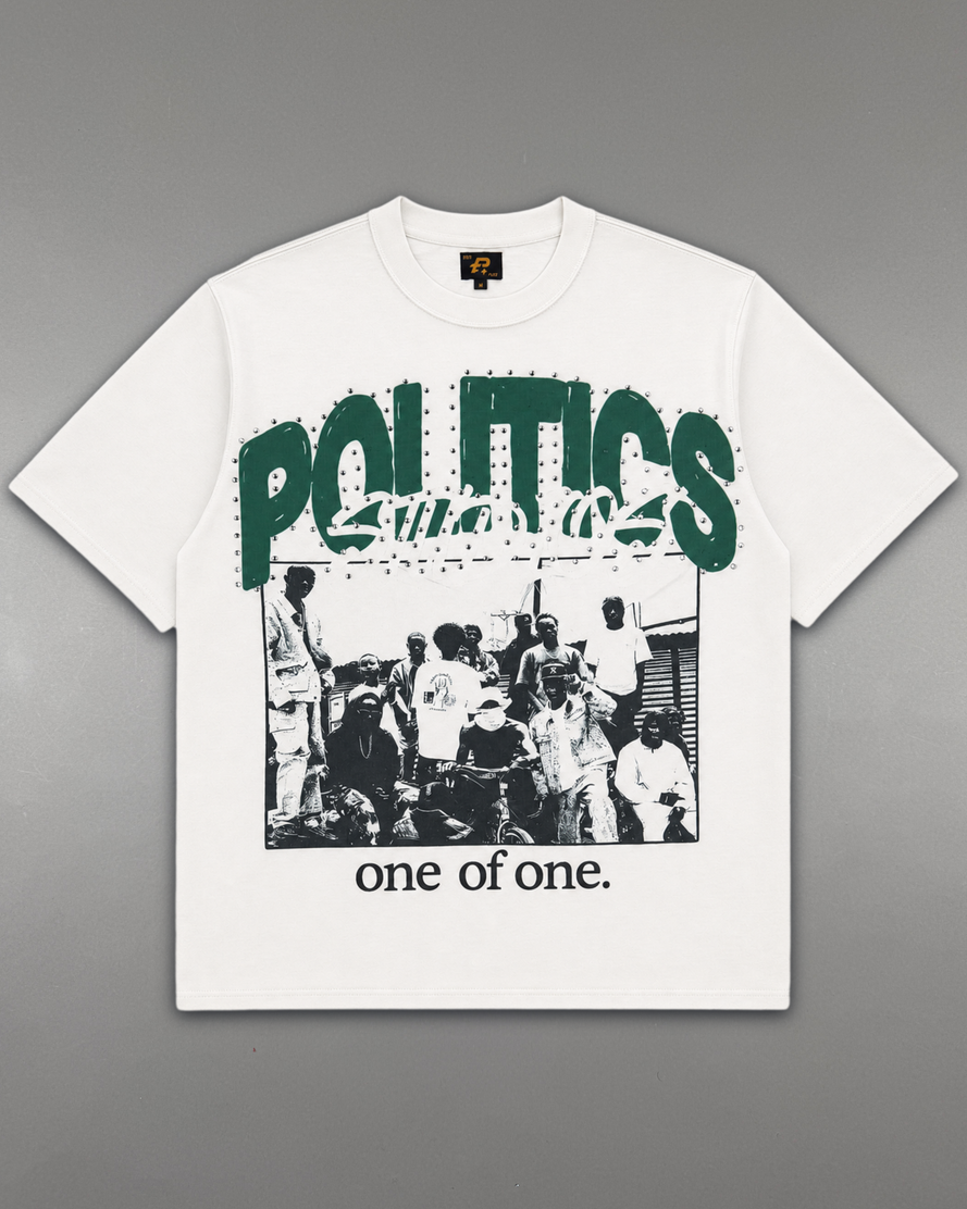 A white short-sleeve graphic t-shirt laid flat on a gray background. The shirt features a large green 'POLITICS' text graphic across the chest, a black-and-white photograph of a crowd underneath, and the phrase 'one of one.