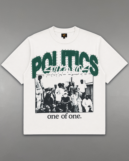 A white short-sleeve graphic t-shirt laid flat on a gray background. The shirt features a large green 'POLITICS' text graphic across the chest, a black-and-white photograph of a crowd underneath, and the phrase 'one of one.