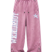 A pair of distressed, faded pink sweatpants featuring a vertical 'WKNDRIOT' graphic on the left leg, a light blue star on the right thigh, and metal snap details along the side pockets and cuffs.