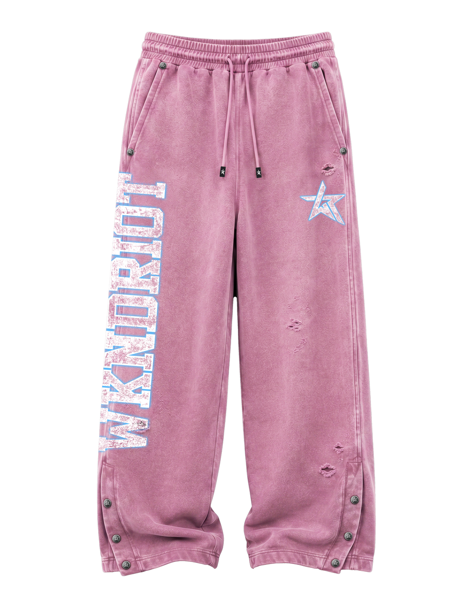 A pair of distressed, faded pink sweatpants featuring a vertical 'WKNDRIOT' graphic on the left leg, a light blue star on the right thigh, and metal snap details along the side pockets and cuffs.