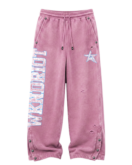 A pair of distressed, faded pink sweatpants featuring a vertical 'WKNDRIOT' graphic on the left leg, a light blue star on the right thigh, and metal snap details along the side pockets and cuffs.