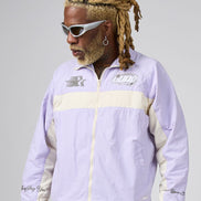 A man with blonde dreadlocks and white sunglasses wearing a light purple and cream windbreaker jacket, photographed against a neutral gray studio background.