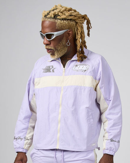 A man with blonde dreadlocks and white sunglasses wearing a light purple and cream windbreaker jacket, photographed against a neutral gray studio background.