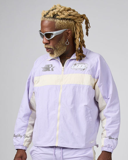 A man with blonde dreadlocks and white sunglasses wearing a light purple and cream windbreaker jacket, photographed against a neutral gray studio background.