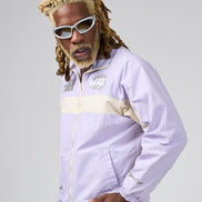 A man with blonde dreadlocks and white sunglasses wearing a light purple and beige windbreaker jacket, posed against a solid gray studio background.