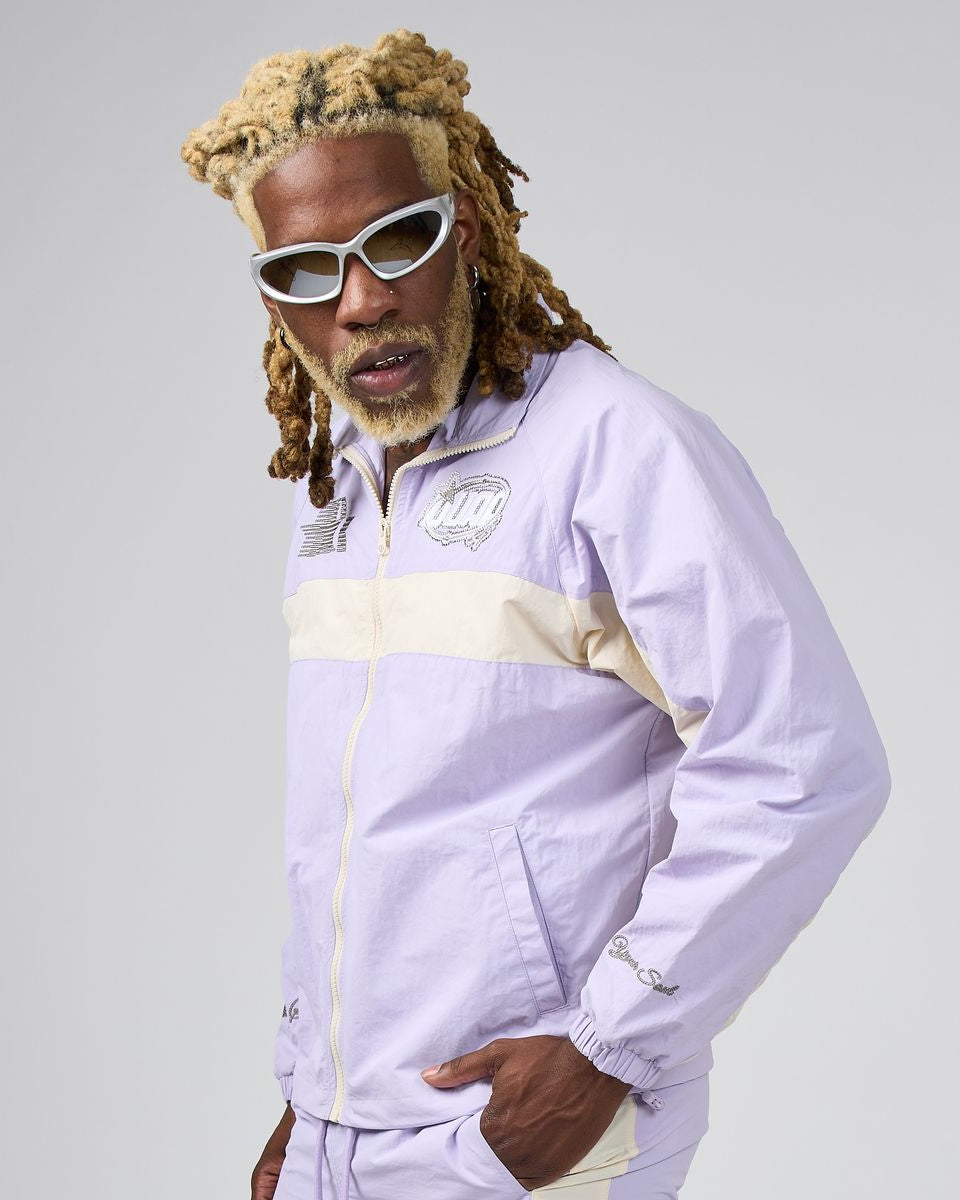 A man with blonde dreadlocks and white sunglasses wearing a light purple and beige windbreaker jacket, posed against a solid gray studio background.