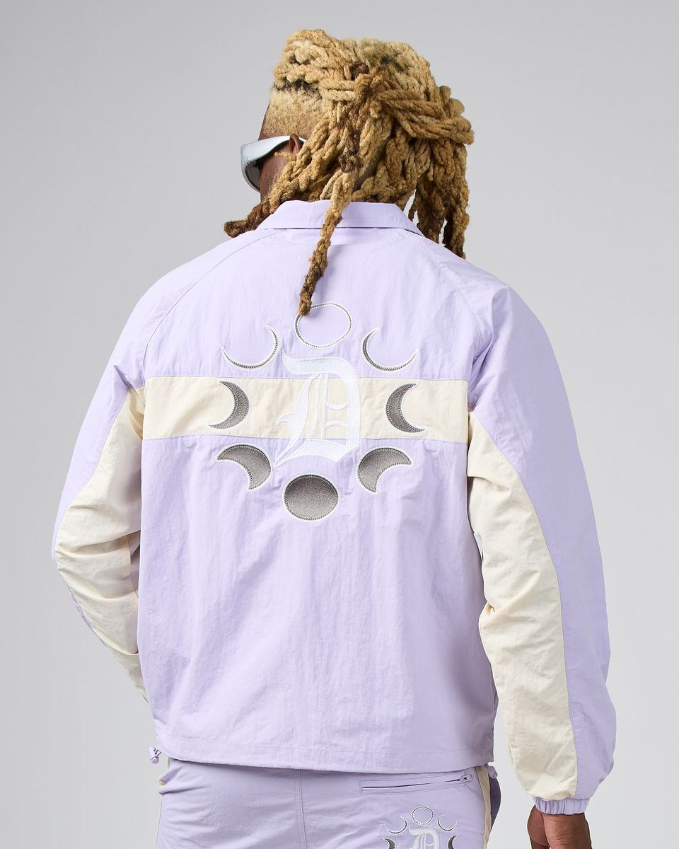 A person shown from the back wearing a light lavender windbreaker jacket with cream-colored sleeves and a moon-phase cutout design across the upper back. The jacket features a large 'JP' monogram logo in the center.