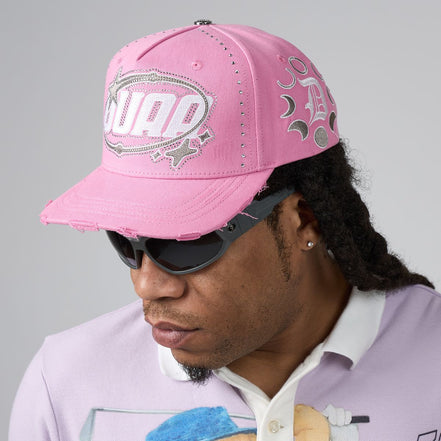 A man wearing a pink distressed baseball cap with 'DUAA' in silver rhinestone lettering on the front, styled with black sunglasses and a light purple polo shirt against a neutral gray background.