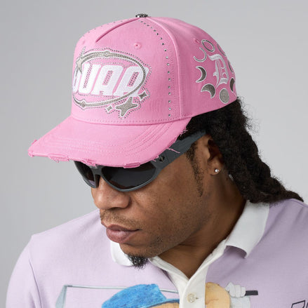A man wearing a pink distressed baseball cap with 'DUAA' in silver rhinestone lettering on the front, styled with black sunglasses and a light purple polo shirt against a neutral gray background.