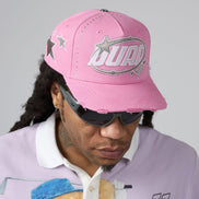A close-up of a person wearing a pink distressed baseball cap with silver rhinestone 'DUAA' lettering and star embellishments, along with dark sunglasses and a light purple polo shirt against a plain gray background.