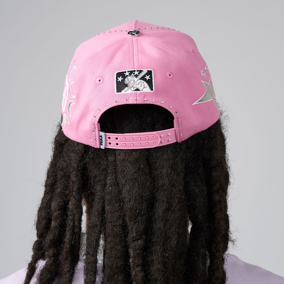 A pink baseball cap shown from the back, worn by a person with long dark dreadlocks. The cap features a black-and-white graphic patch in the center, silver studs, and white graphic details on the sides, set against a plain gray background.