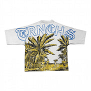 A white short-sleeve t-shirt featuring a large graphic print on the back. The design includes the word 'TRNCHS' in blue outlined letters across the top, above a yellow and black illustrated scene of palm trees and people walking outdoors.