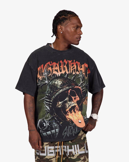 A man wearing a black Sublimation brand graphic t-shirt with a skull and mechanical design, paired with camouflage pants, against a light gray background.