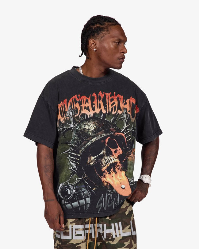 A man wearing a black Sublimation brand graphic t-shirt with a skull and mechanical design, paired with camouflage pants, against a light gray background.