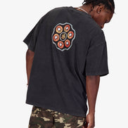 A man wearing a faded black oversized t-shirt with a colorful circular graphic on the back, paired with camouflage pants, photographed against a light gray background.