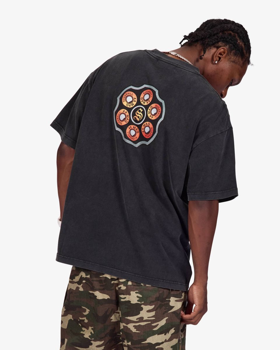 A man wearing a faded black oversized t-shirt with a colorful circular graphic on the back, paired with camouflage pants, photographed against a light gray background.