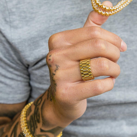 Hand wearing a gold chain bracelet and wearing the gold presidential ring with a gray shirt background