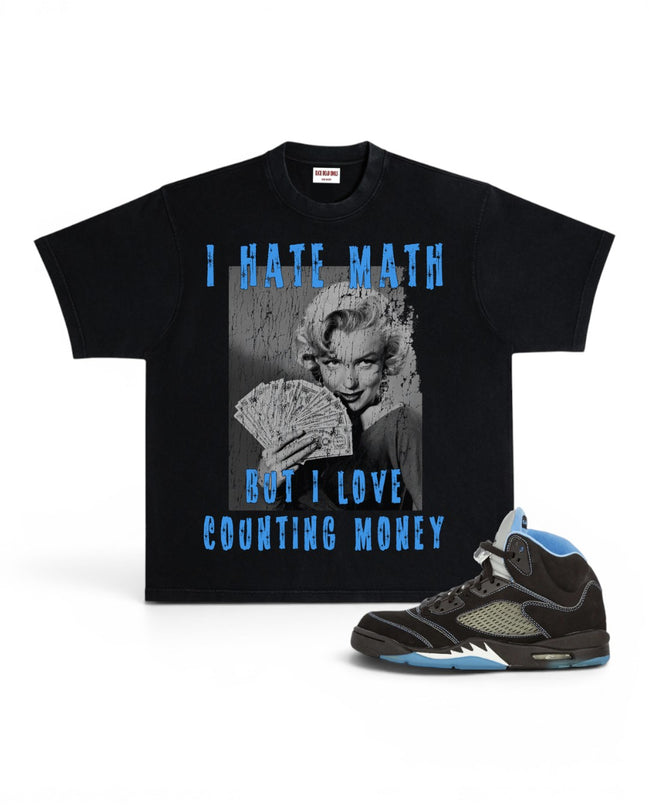 A black graphic t-shirt featuring a Marilyn Monroe print with the text 'I HATE MATH BUT I LOVE COUNTING MONEY' in distressed blue lettering, shown alongside a black and light blue sneaker on a white background.