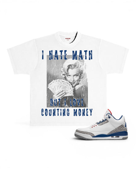 A white graphic t-shirt featuring a black-and-white photo of Marilyn Monroe holding cards with blue text reading 'I HATE MATH BUT I LOVE COUNTING MONEY', shown next to a white sneaker with blue and gray accents on a white background.