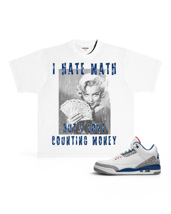 A white graphic t-shirt featuring a black-and-white photo of Marilyn Monroe holding cards with blue text reading 'I HATE MATH BUT I LOVE COUNTING MONEY', shown next to a white sneaker with blue and gray accents on a white background.