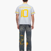 Person wearing a white sports jersey with yellow numbers and text, paired with blue jeans featuring gold designs.