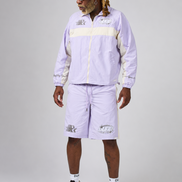 A model wearing a light purple and cream tracksuit consisting of a zip-up jacket and matching shorts, styled with white socks and beige-and-black sneakers against a neutral gray studio background.